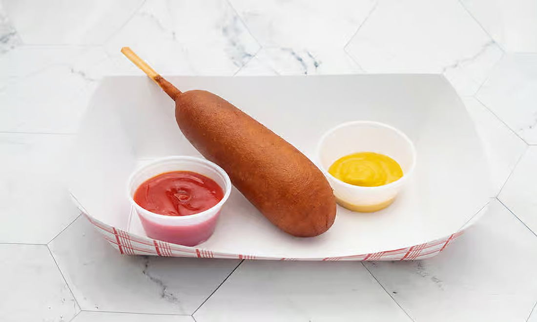 A golden corn dog on a paper tray with ketchup and mustard cups