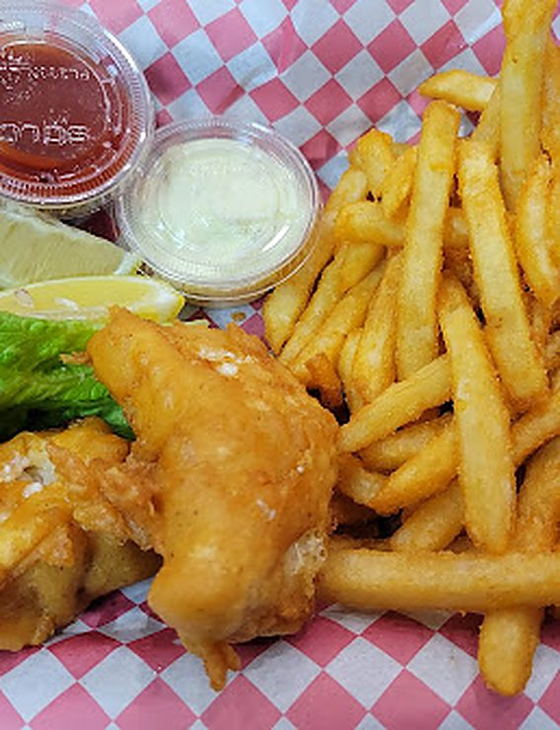 Golden battered fish and crispy fries in a red-checked basket with tartar sauce, cocktail sauce and a lemon wedge